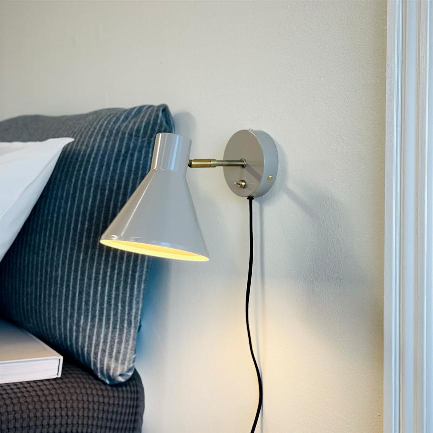 Væglampe: en grå, konisk lampe lyser nedad; monteret ved en stribet blå pude og sengens hovedgærde, med sort ledning hængende langs en lys væg.