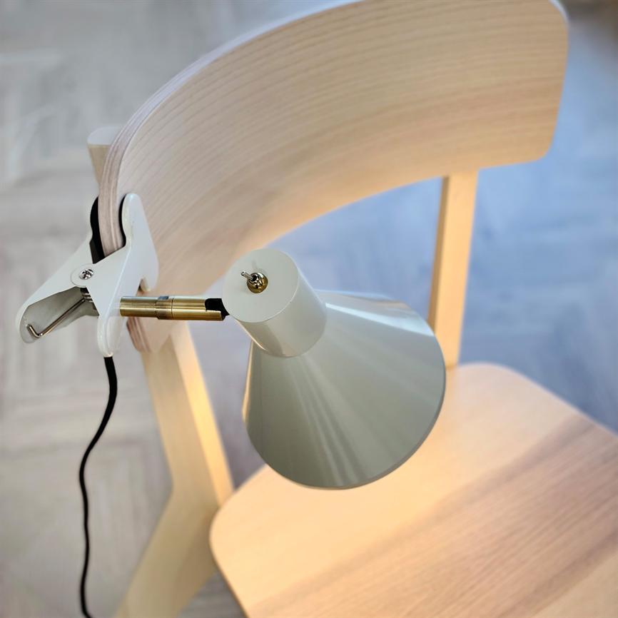 Klemme-lampe fastgjort på ryglænet af en lys træstol, lyser nedad og kaster varmt lys over sædet; sort ledning hænger ned; rum med lyst trægulv i baggrunden.