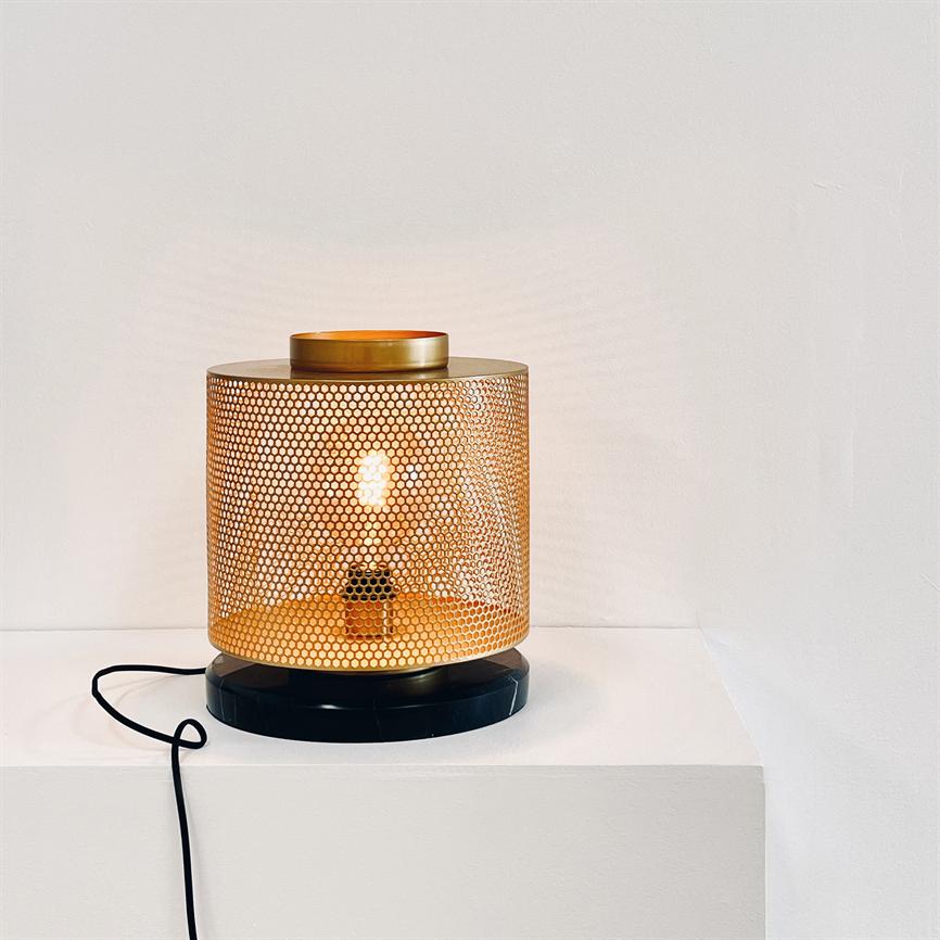 En cylindrisk guldfarvet bordlampe afgiver varmt, mønstret lys gennem en perforeret metalskærm; den står på en hvid sokkel foran en lys væg, med en sort ledning ved basen.