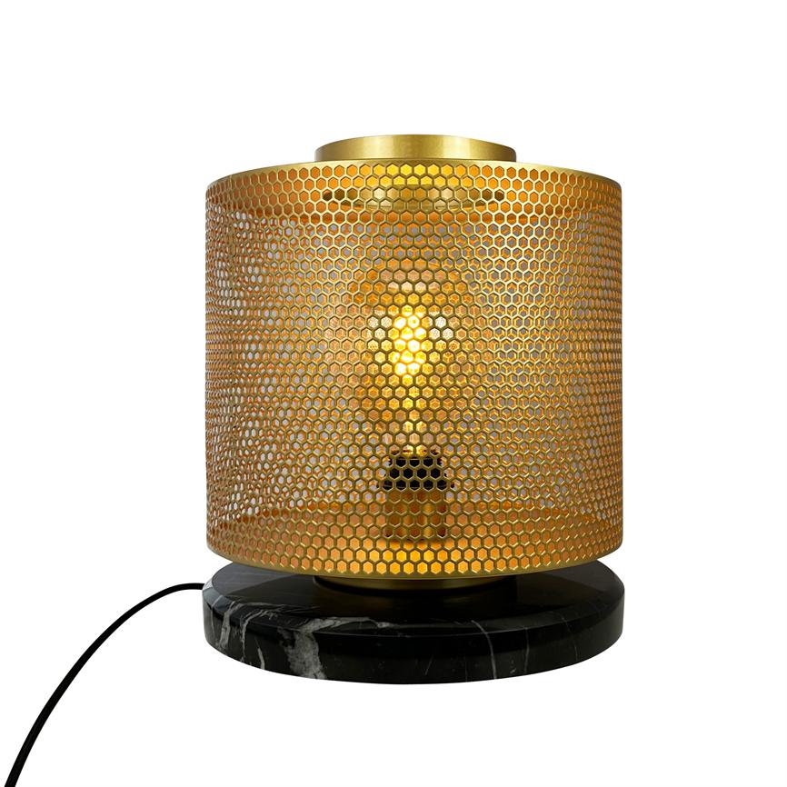 Guldbelyst bordlampe med perforeret honningkagelignende cylinderafskærmning, hvor en tændt pære skinner gennem mønsteret; står på rund sort marmorbase med synlig sort ledning på hvid baggrund.