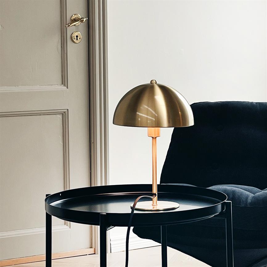 Messing bordlampe står tændt på et rundt sort sidebord; dens varme skær falder over en mørk fløjlsstol og en detaljeret hvid dør i et lyst, minimalistisk opholdsrum.