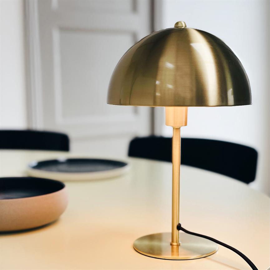 Messingbordlampe kaster varmt, afdæmpet lys over et lyst bord; den står på en rund fod med sort ledning, omgivet af sorte stole og to keramikfade i et minimalistisk rum.