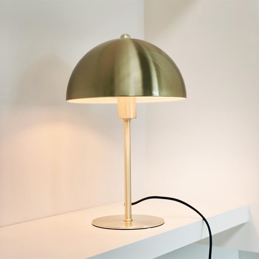 Bordlampe med rund grøn-guld kuppel skærm står tændt på en hvid hylde; slank messingstang og rund fod, sort ledning løber ud mod højre foran en lys væg.