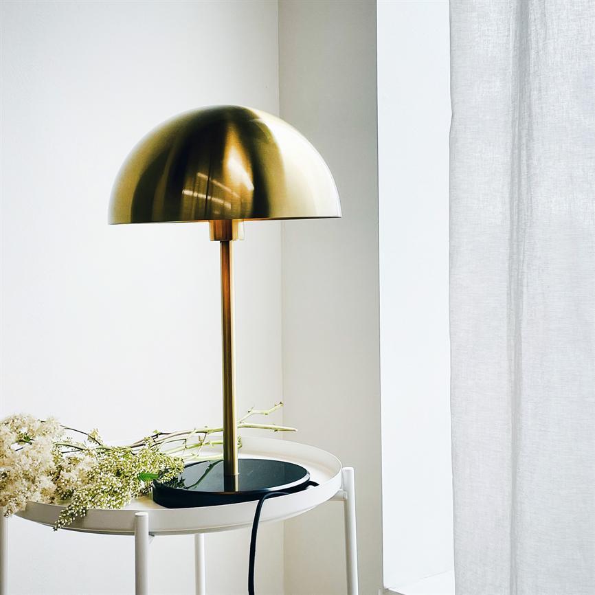 En guldfarvet kuppelformet bordlampe står på et rundt hvidt sidebord; sort base og ledning, hvide blomster ligger ved siden af, ved et lyst vindue med en hvid gardin.