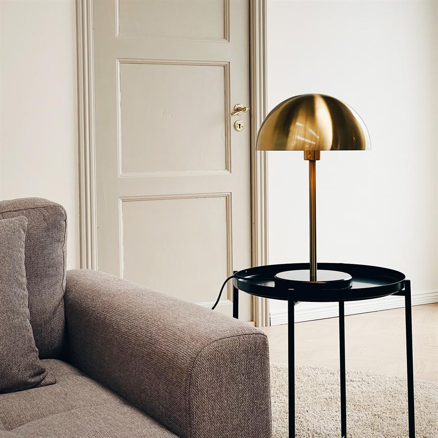 En messingkuppelformet bordlampe står på et rundt sort sidebord ved siden af en grå sofa i en lys, minimalistisk stue med fyldør, lyst trægulv og lyst tæppe.