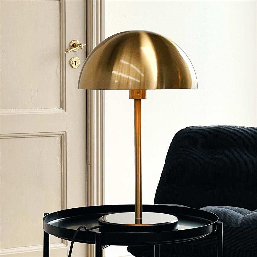 En rund messinglampe står på et sort rundt sidebord; dens kuppelformede skærm hviler på en slank messingstang. Baggrunden er en beige dør og en mørk, polstret lænestol.