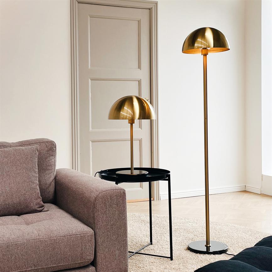 To guldlamper står ved en grå sofa; den ene på et sort sidebord, den anden som gulvlampe på trægulvet, omgivet af lyse vægge, en beige dør og et lyst tæppe.