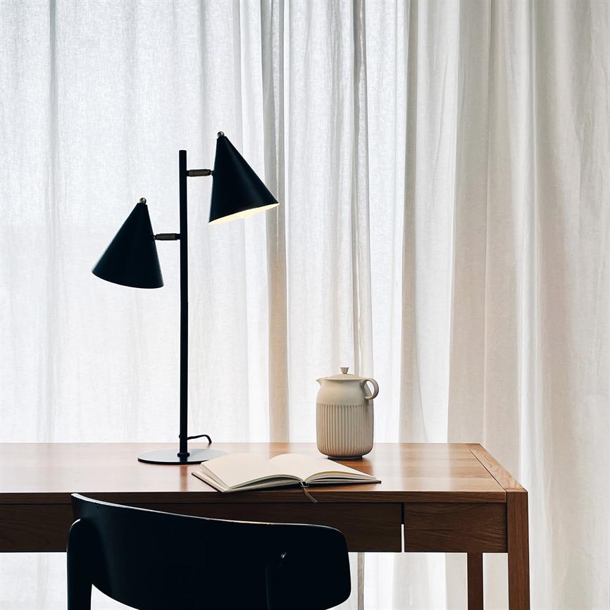 En sort dobbeltkeglelampe peger ned mod bordet; på træbordet ligger en åben notesbog og en keramisk kande, en sort stol foran og bløde hvide gardiner i baggrunden.