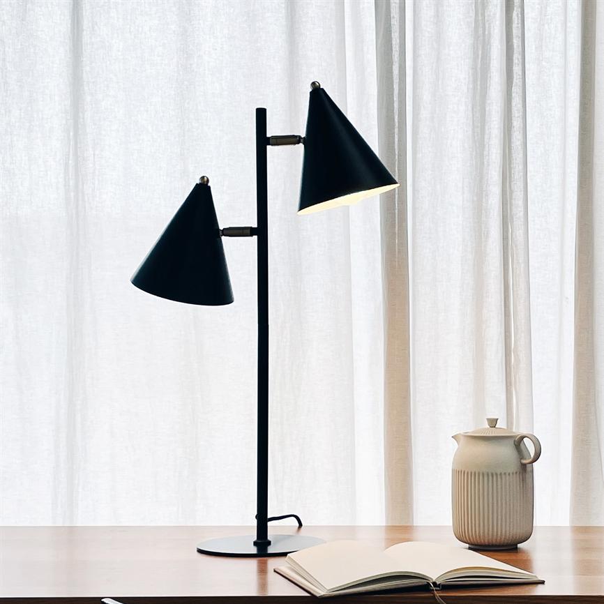 Sort dobbelt-konisk bordlampe med en tændt skærm kaster dæmpet lys over en åben notesbog på et træbord ved siden af en keramisk kande foran lyse gardiner.
