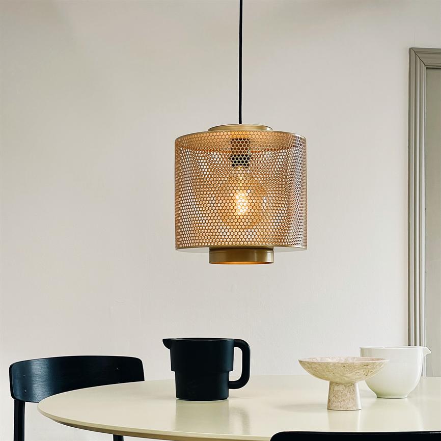 En cylindrisk perforeret messingpendel lyser varmt, hængende over et rundt lyst bord med en sort kande, en marmoragtig skål og en hvid skål i et lyst, minimalistisk rum.