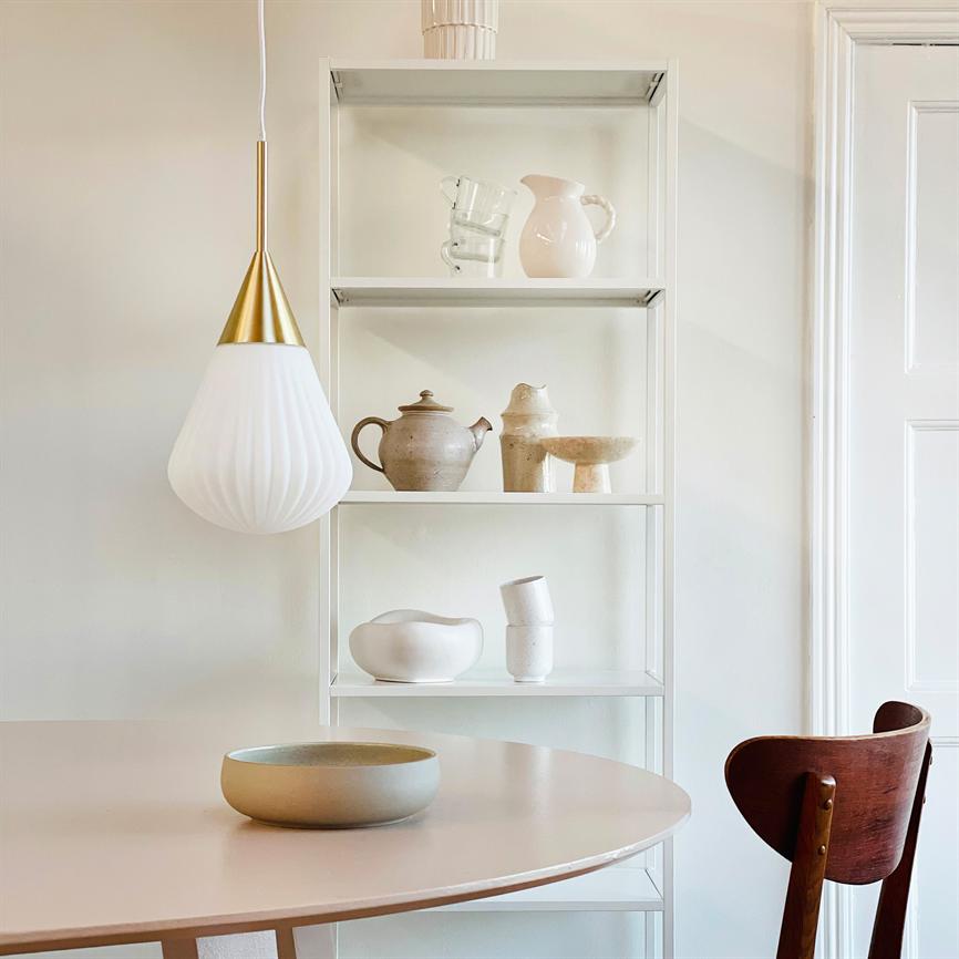 Hvid-guld pendel hænger over et rundt, lyserødt bord; en beige keramisk skål står midt på bordet. En hvid reol i baggrunden viser neutrale keramikvaser og skåle; en mørk træstol ses til højre.