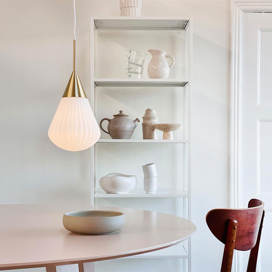 En hvid-guld pendel lyser blødt over et rundt bord; bagved står en åben hvid reol med beige keramikvaser, og en mørk træstol ses til højre.