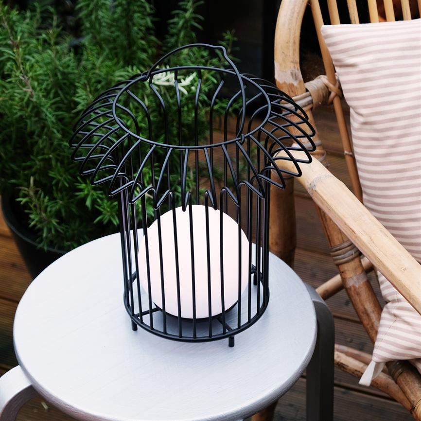 Sort metallanterne med en oplyst hvid glaskugle står på et lille rundt gråt bord; ved siden af en rattanstol med stribet pude og en pottet grøn plante i baggrunden.