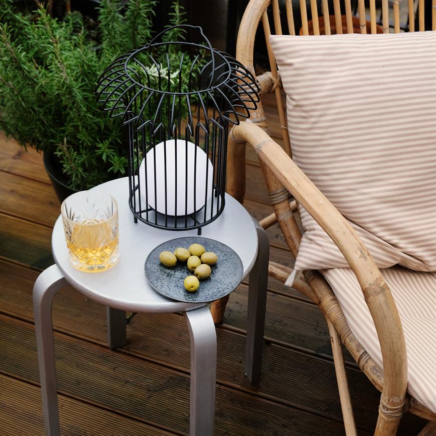 Lille rundt sidebord bærer en sort metal-lanterne med kuglelys, et whiskyglas og en tallerken med grønne oliven; ved siden af står en flettet rattanstol med stribet pude på en træterrasse.