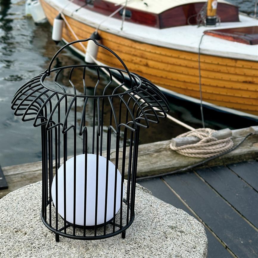 Sort metallanterne i burform står på en granitklump og rummer en hvid kuglelampe; i baggrunden ligger en gulbrun træbåd fortøjet ved en træbro med snoet tov.