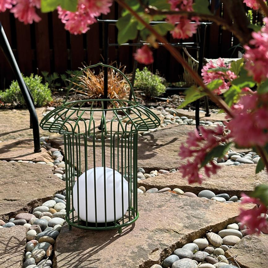 En grøn metallygte formet som et bur rummer en hvid kugle, står på flade sten mellem små runde sten på en solbeskinnet stenbelægning, omgivet af lyserøde blomster.