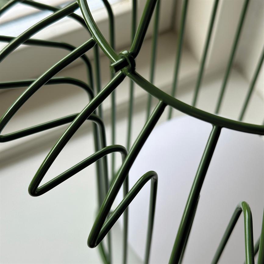 Grøn metaltrådkurv danner bladlignende, buede tråde og står ved en vindueskarm i blødt dagslys med en neutral, udvisket baggrund.