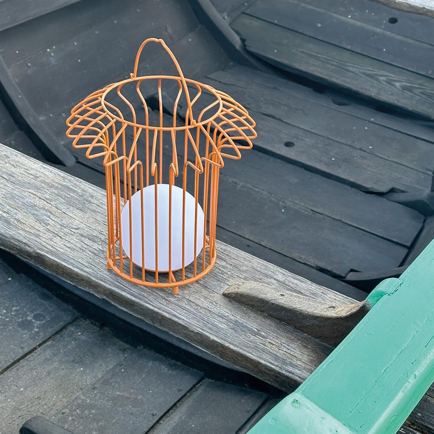 Orange trådbur-lanterne med hvid kuglelyskilde står på et slidt træbræt i en sort robåd, omgivet af sorte sæder og en grønmalet ræling.