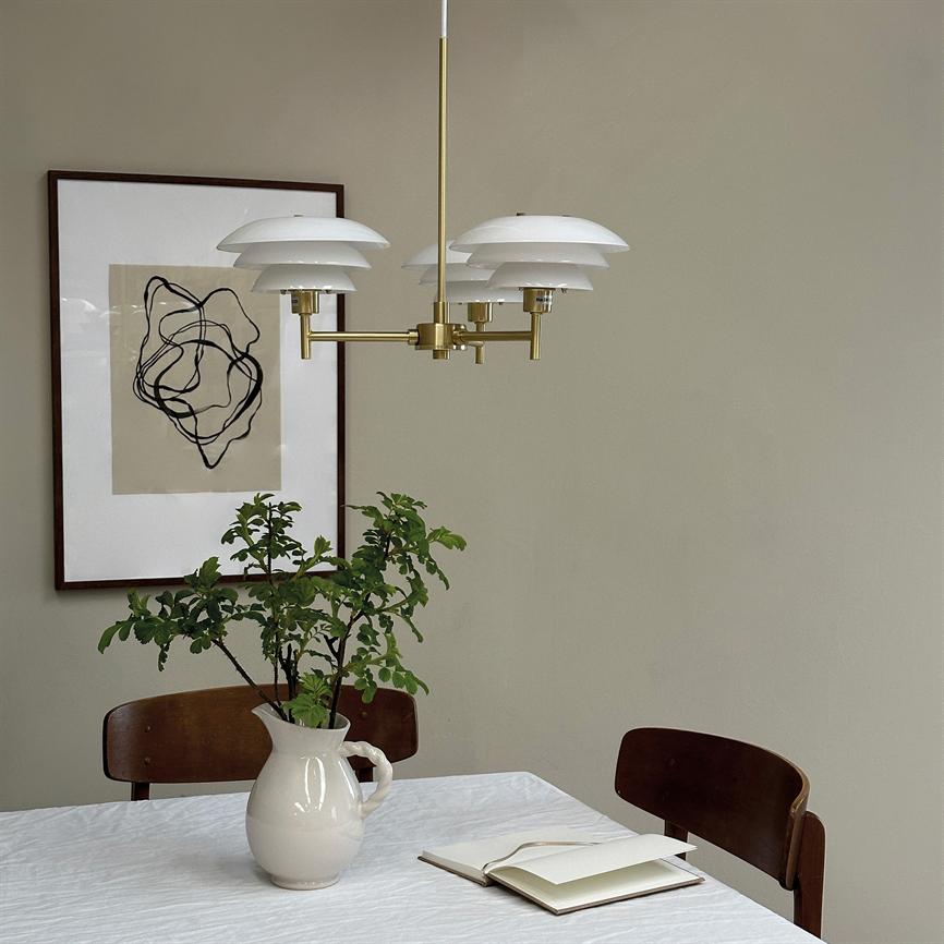 Pendel med hvide skærme hænger over et hvidt dugdækket bord; en keramikkande med grønne grene står på bordet, en åben notesbog ligger ved en træstol, beige væg med abstrakt kunst.