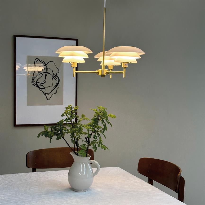 Loftlampe hænger og lyser blødt over et spisebord; en hvid kande med grønne grene står på hvid dug mellem to brune stole foran en grå væg med abstrakt billede.