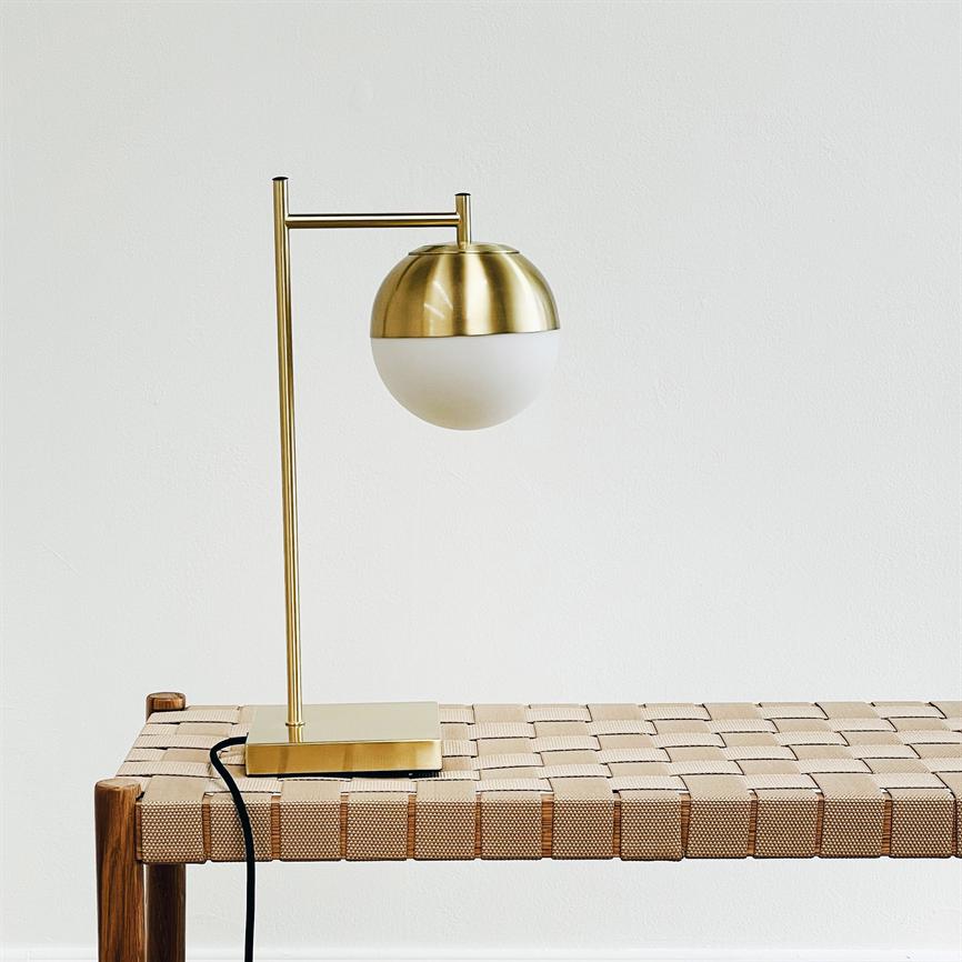 Guldfarvet bordlampe med halvkugleformet opalglaskærm står på en vævet træbænk; arm holder kugleskærmen ud fra basen i et lyst, minimalistisk rum, med synlig sort ledning.