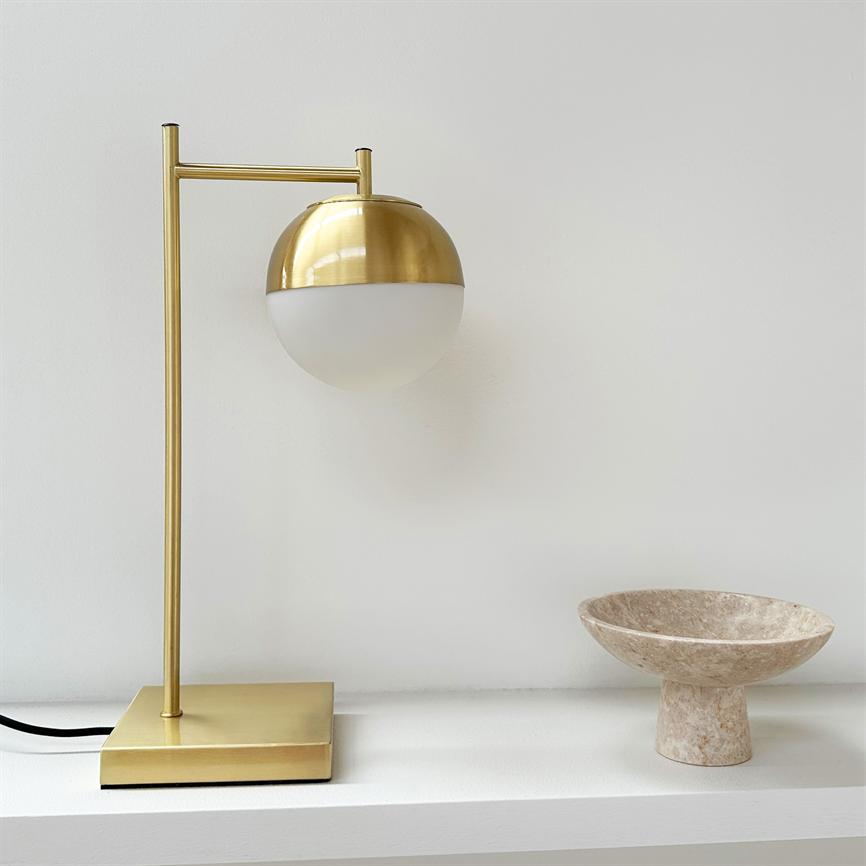 Guldfarvet bordlampe med halvkugleformet opalglas er monteret på en slank stang og står på en firkantet base på en hvid hylde; til højre en beige marmorskål.