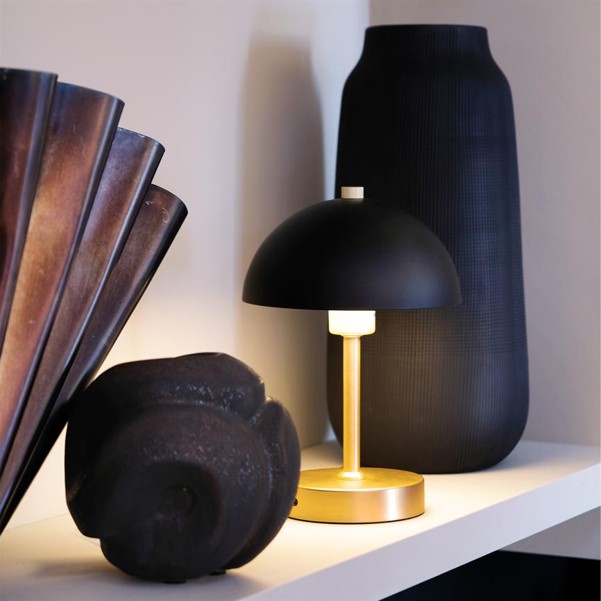 En lille bordlampe med sort kuppel og guldfarvet fod afgiver varmt, blødt lys; den står på en hylde mellem en mørk vase og en sort skulpturel sten foran en lys væg.
