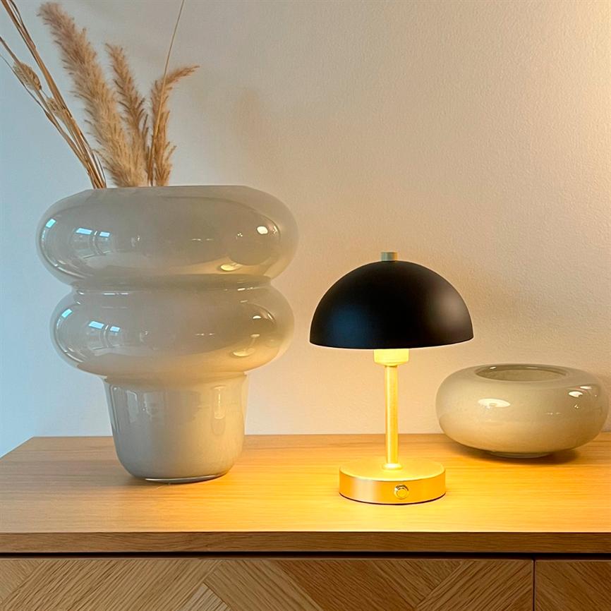 Stor blank keramikkrukke med tørrede pampasgræsser, en lille sort og messingfarvet bordlampe som lyser varmt, og en lav rund vase på et træskab foran en neutral væg.