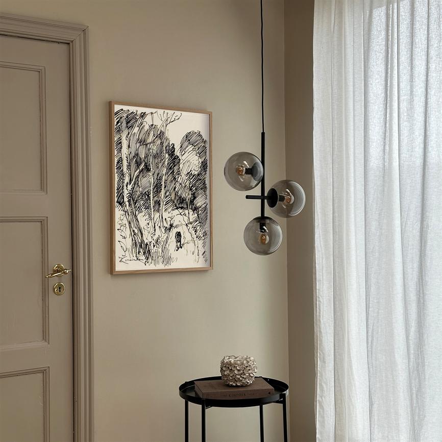 En indrammet sort-hvid skitse af træer hænger på en beige væg; en tre-globet glaspendel hænger ved siden af, og et lille rundt sidebord med bog og dekorativ genstand står ved et lyst gardin.
 Tekst: KINFOLK