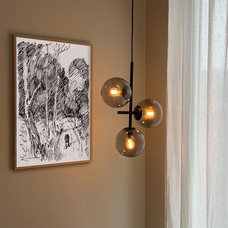 En moderne loftslampe med tre røgfarvede glaskupler, der udsender varmt glødelampe-lys; ved siden af en indrammet sort-hvid skitse hænger på beige væg, lyse gardiner til højre.