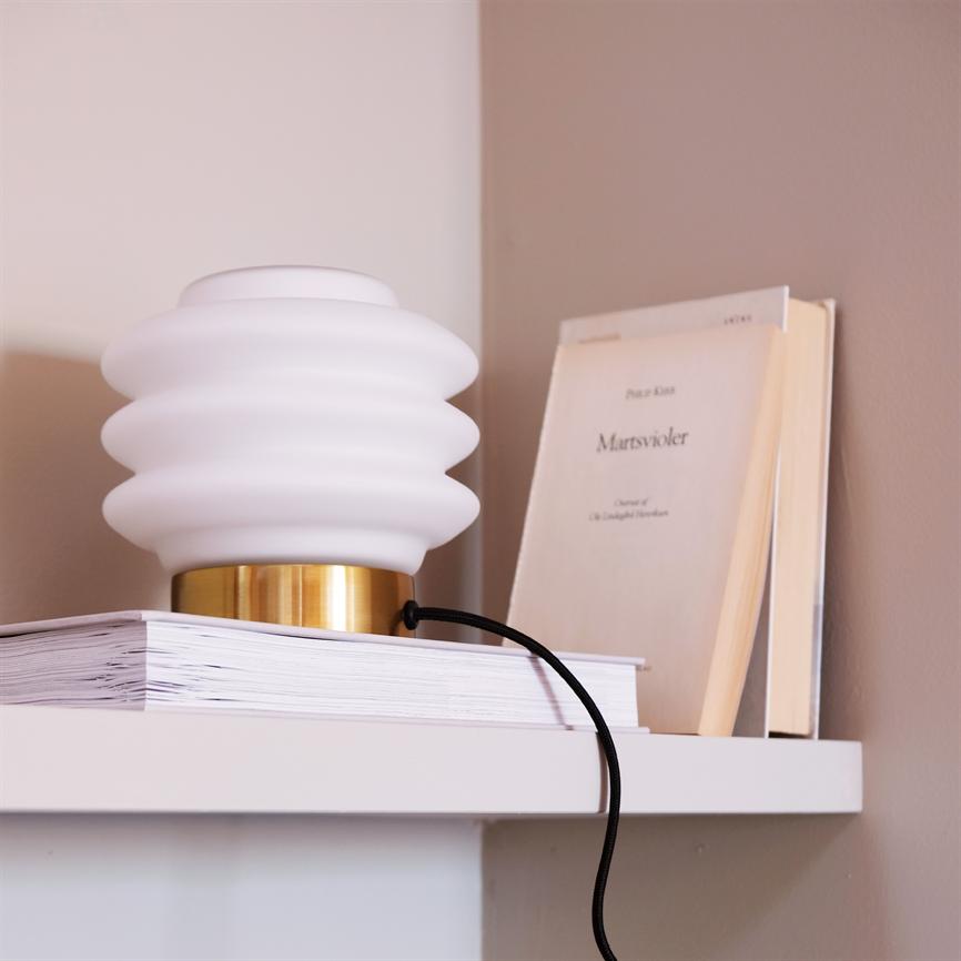 En hvid, bølget bordlampe står på en stak bøger på en hvid hylde; en beige bog står opret bagved mod en lysebrun væg. 
 Tekst på bogomslaget: "Martsvioler" (øvrig tekst ikke tydelig).