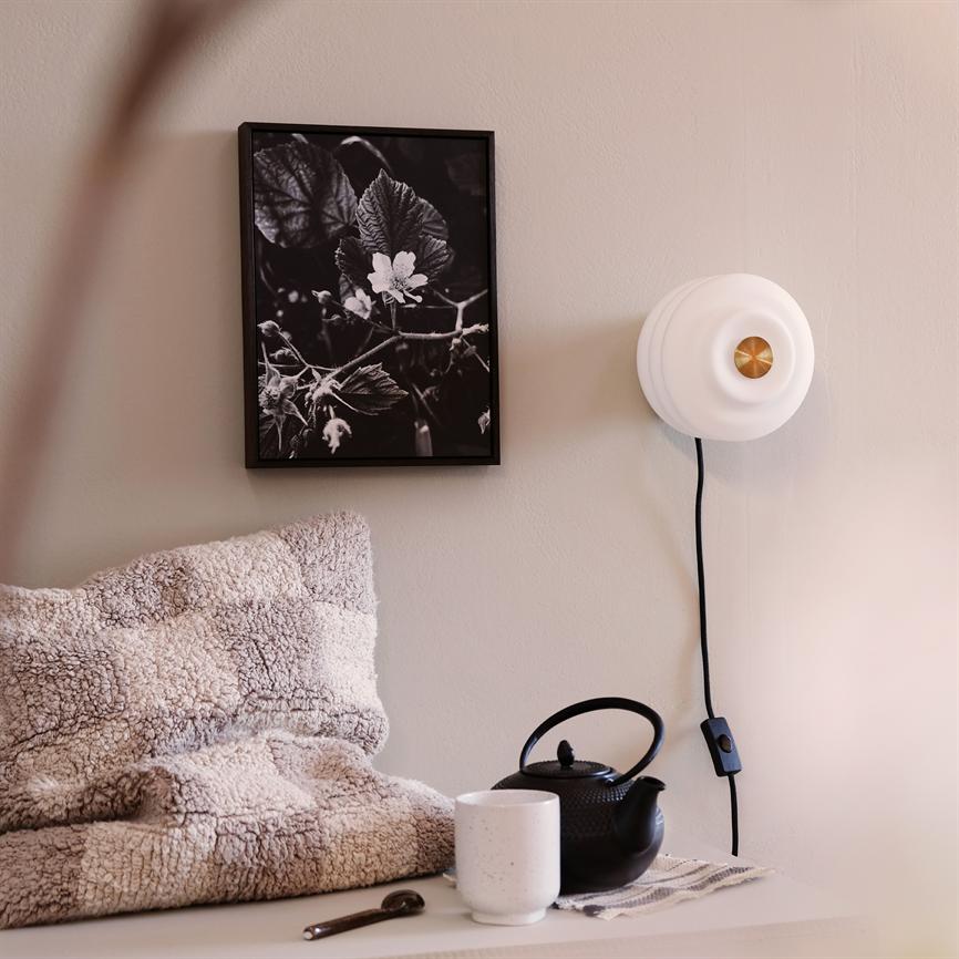 Et indrammet sort-hvidt blomsterfotografi hænger stille på væggen, flankeret af en hvid væglampe; foran ligger en blød plaid, en sort tekande og en hvid kop på en hylde.