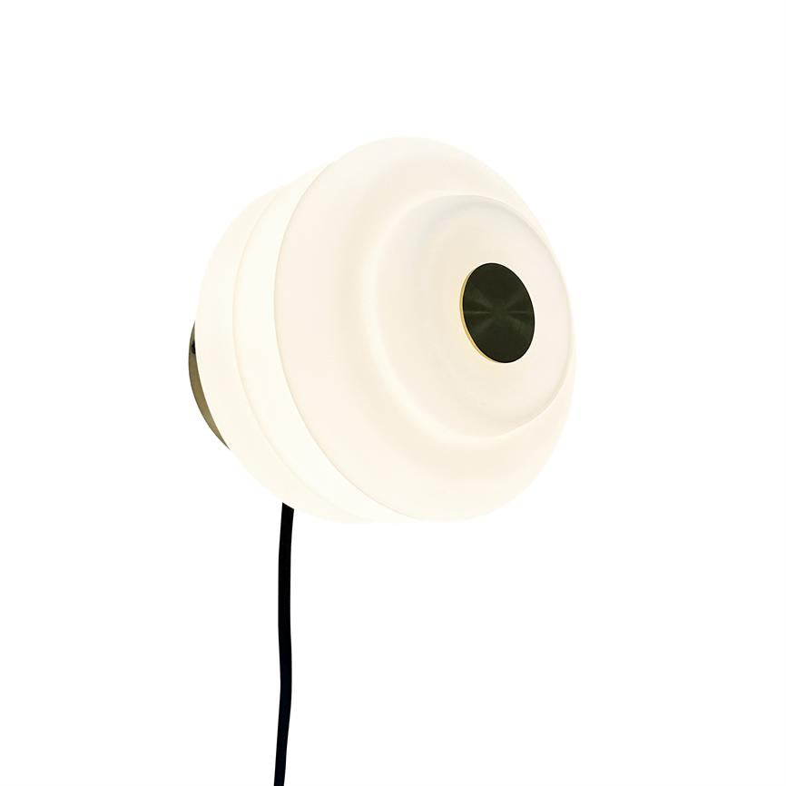 Rund væglampe lyser svagt; lagdelt hvid skærm med en mørk, metallisk cirkel i midten. Sort ledning hænger ned fra lampen, monteret mod en neutral hvid baggrund.