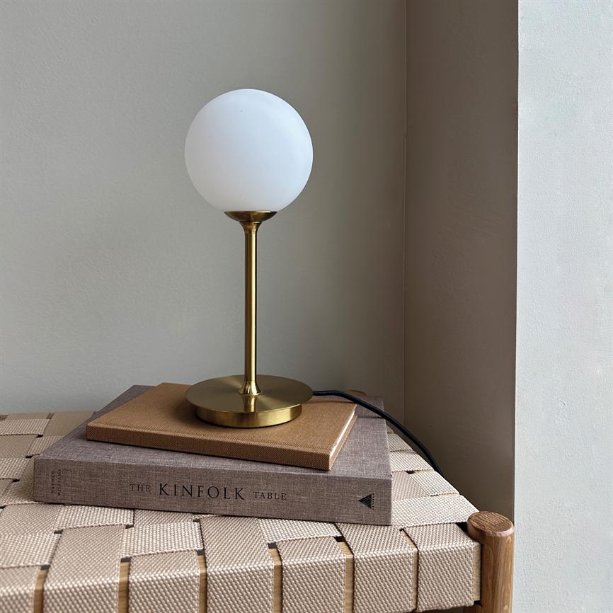 Bordlampe med rund hvid glasskærm og messingstativ står på to bøger på en flettet bænk i et lyst væghjørne. 
 Tekst på bogryg: "THE KINFOLK TABLE".