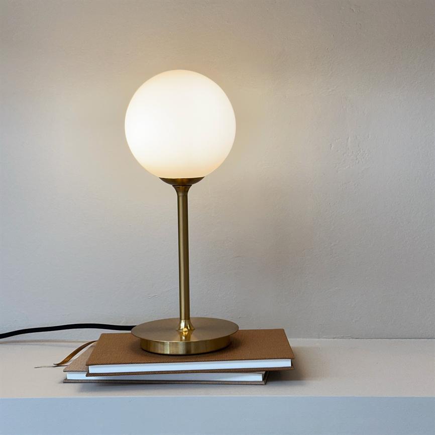 Bordlampe med rund opalglaskuppel står og lyser på en messingfod oven på to notesbøger på en hvid hylde foran en lys, let struktureret væg.