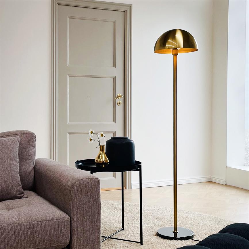 Gulvlampe i messing står tændt ved et lille sort sidebord; bordet har en sort vase og en lille gylden vase med hvide blomster, foran en beige sofa og lukket dør.