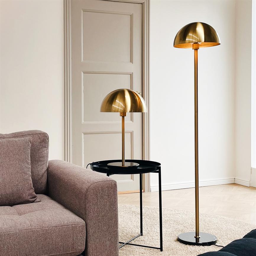To lamper i børstet messing — en bordlampe på et rundt sort sidebord og en høj gulvlampe — står og kaster varmt lys ved en beige sofa på et lyst tæppe foran en dør.