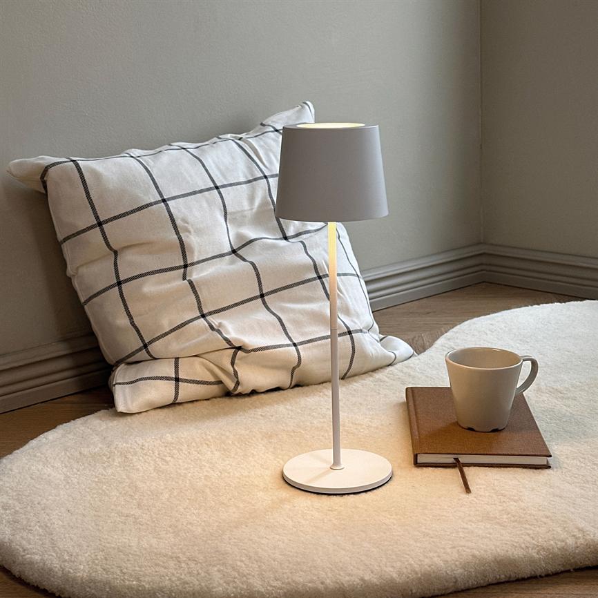 Bordlampe står tændt på et rundt, lyst uldmåtte; ved siden ligger en ternet pude lænet mod væggen og en notesbog med krus på trægulvet i et roligt hjørne.