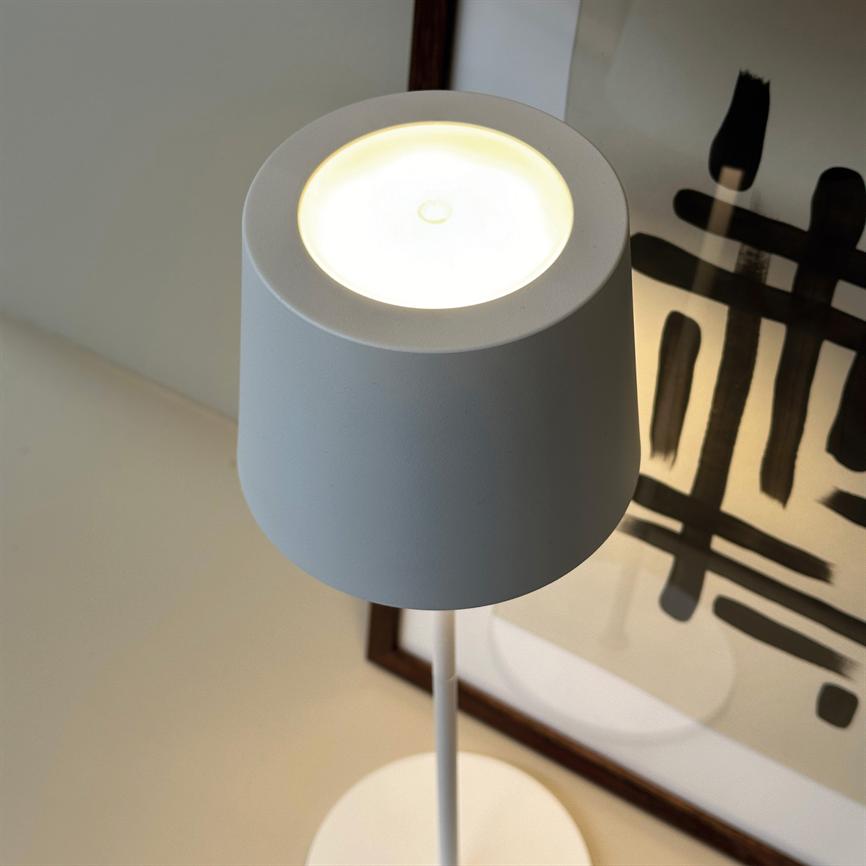 En hvid bordlampe står tændt, dens koniske skærm kaster blødt lys nedad, placeret på et bord foran en indrammet plakat med sorte penselstrøg.