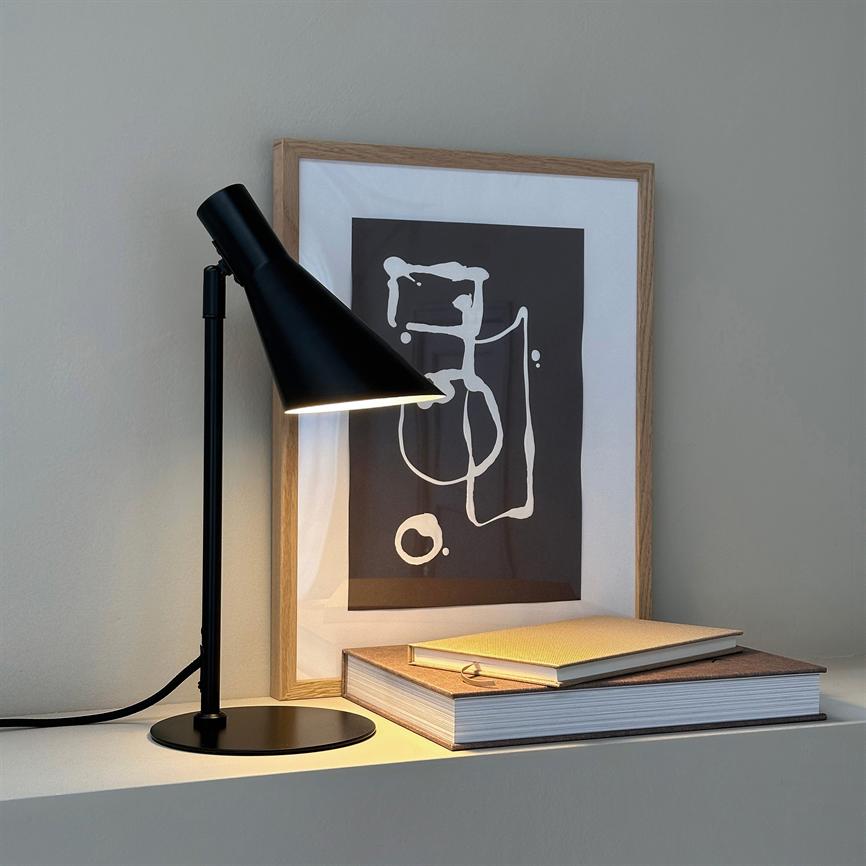 En sort bordlampe lyser ned på en stak bøger og et indrammet abstrakt motiv. Genstandene står på en hvid hylde foran en neutral, grå væg.