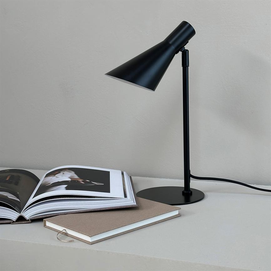 Sort metalbordlampe peger nedad over en åben fotobog; en lukket notesbog ligger ved siden af på en lys bordplade med en synlig ledning foran en lysegrå væg.