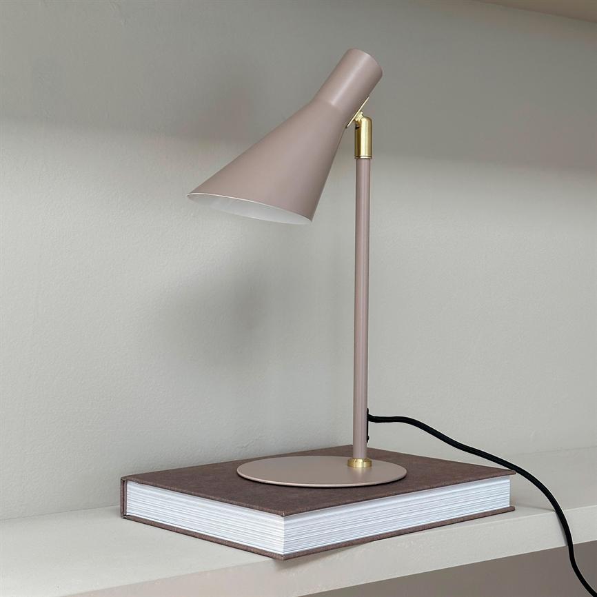 Bordlampe peger skråt nedad, står på en lukket, tyk bog på en hylde foran en lysgrå væg; metalstang og skærm i støvet rosa med sort ledning.