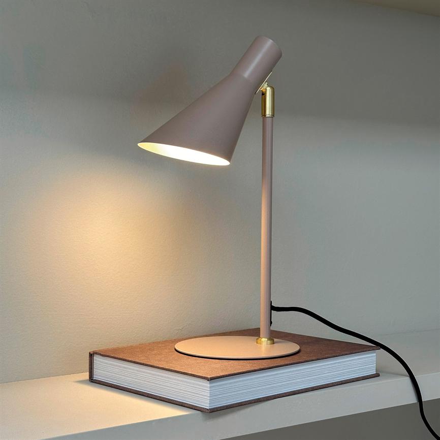 Bordlampe lyser blødt ned på en tyk indbundet bog, placeret på en hylde foran en lys væg; slank støvet rosa lampe med gulddetalje og synlig sort ledning.