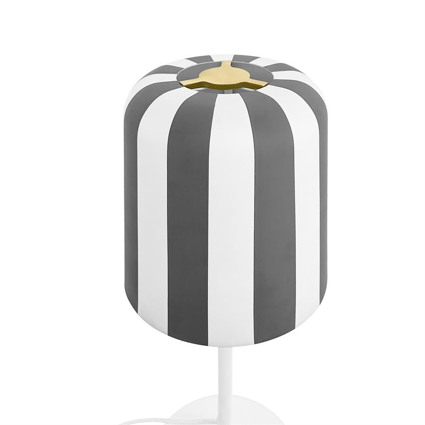 Lampe med cylindrisk skærm i lodrette grå og hvide striber, gylden fatning øverst, monteret på tynd hvid fod foran hvid baggrund.