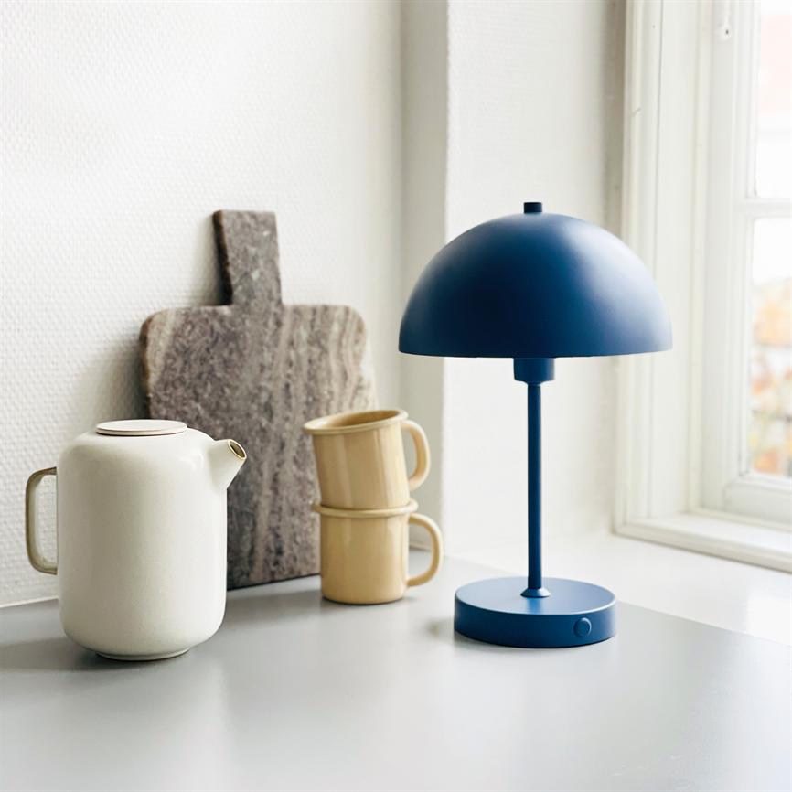 En blå bordlampe står på en lys vindueskarm sammen med en hvid tekande, to stablede beige krus og et marmor-skærebræt i et lyst køkkenrum.