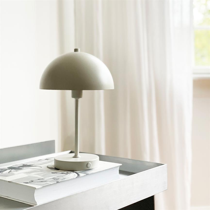 En lille, lys grå bordlampe står på en tyk bog i en metalbakke på et sidebord, foran bløde hvide gardiner og et indrammet vindue.
