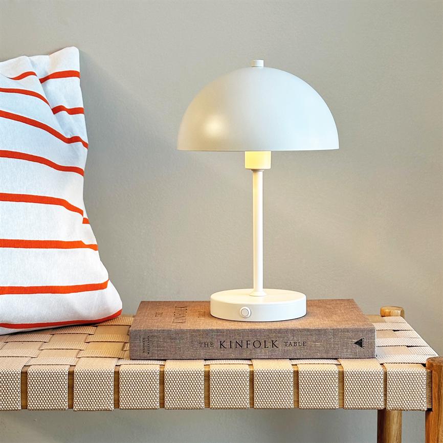 En hvid svampeformet bordlampe står tændt på en indbundet bog på en flettet træbænk; til venstre en hvid pude med orange striber og en neutral væg i baggrunden. Tekst: "THE KINFOLK TABLE".