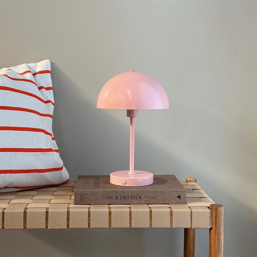 Lyserød bordlampe står på en bog og et flettet træbænk ved en lys væg, ved siden af en hvid pude med røde striber. Tekst: THE KINFOLK TABLE