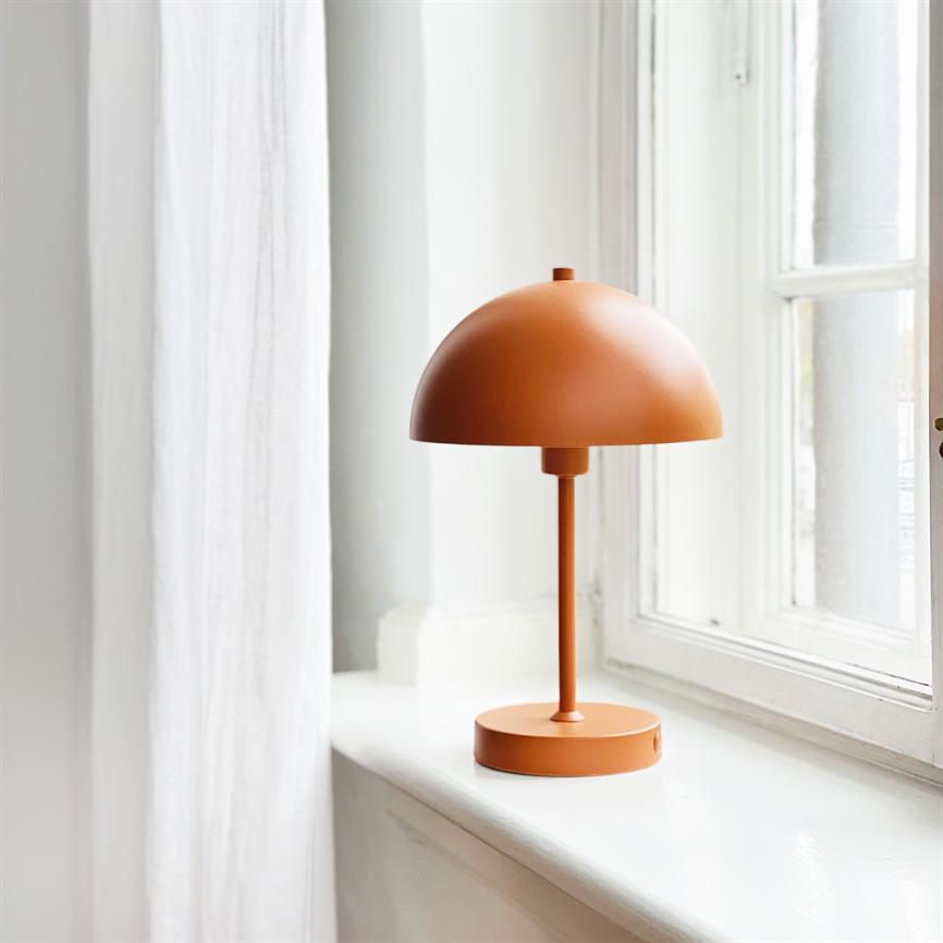 En orange, klokkeformet bordlampe står på en hvid vindueskarm ved et vindue; blødt dagslys strømmer ind, flankeret af et lyst, let gardin.