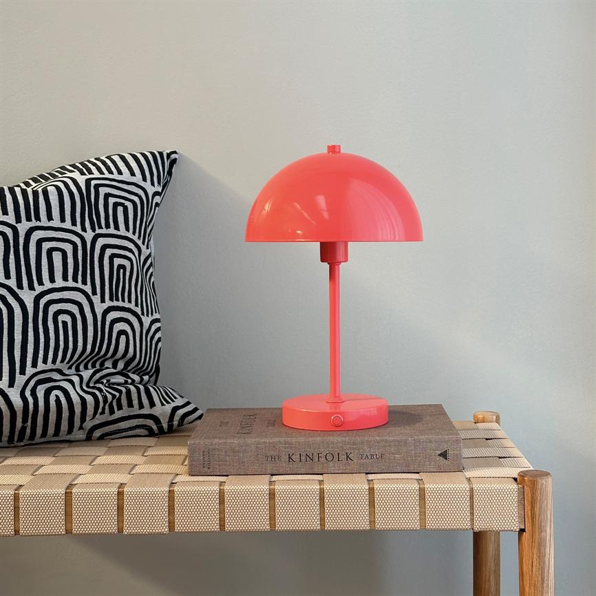 Koralrød bordlampe står på en bog (”THE KINFOLK TABLE”) på en flettet bænk; sort-hvid, geometrisk mønstret pude til venstre; lys, neutral væg i baggrunden.
 
 Tekst: THE KINFOLK TABLE